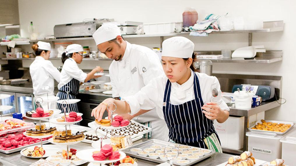 students in chef uniform in a sophisticate kitchen place intricate cakes on platters