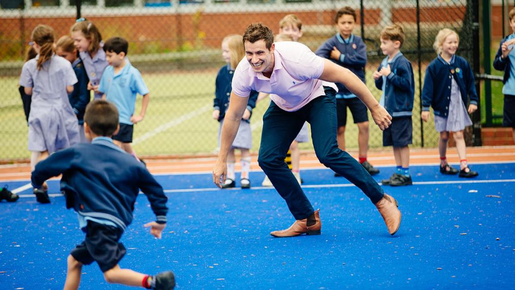 Young white man in a pink polo and jeans, laughing and playing sports games with kids in primary school uniform
