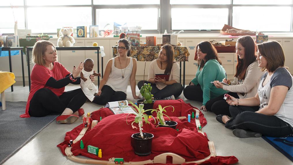 Group of young female students in a daycare model setting, listen to teacher holding a lifelike baby doll