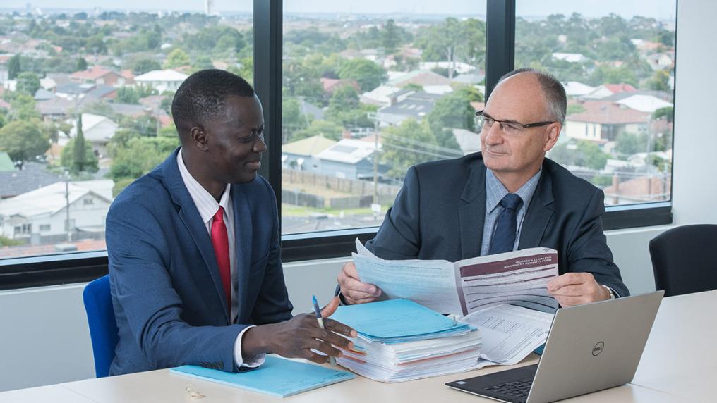 Young African-Australian man in a suit talks to older white man in a suit over papers in a an office with views