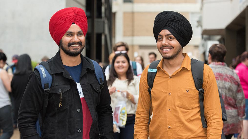 Two Singh students in turbans with backpacks in a crowded courtyard smile at the camera